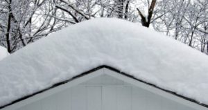 Close up view of roof with a large amount of snow on top. Bare trees in background.